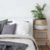 Bed with white and grey linens and a lot of pillows. Piece of interior of the bedroom with a bed, pillows of a headboard and a bedside tube in the form of a stool stairs with a basket and home plant
