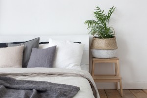 Bed with white and grey linens and a lot of pillows. Piece of interior of the bedroom with a bed, pillows of a headboard and a bedside tube in the form of a stool stairs with a basket and home plant