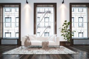 Elegant minimalist apartment living room interior with large windows and a single white couch with cushions and throw rugs on a small carpet over a wood floor.